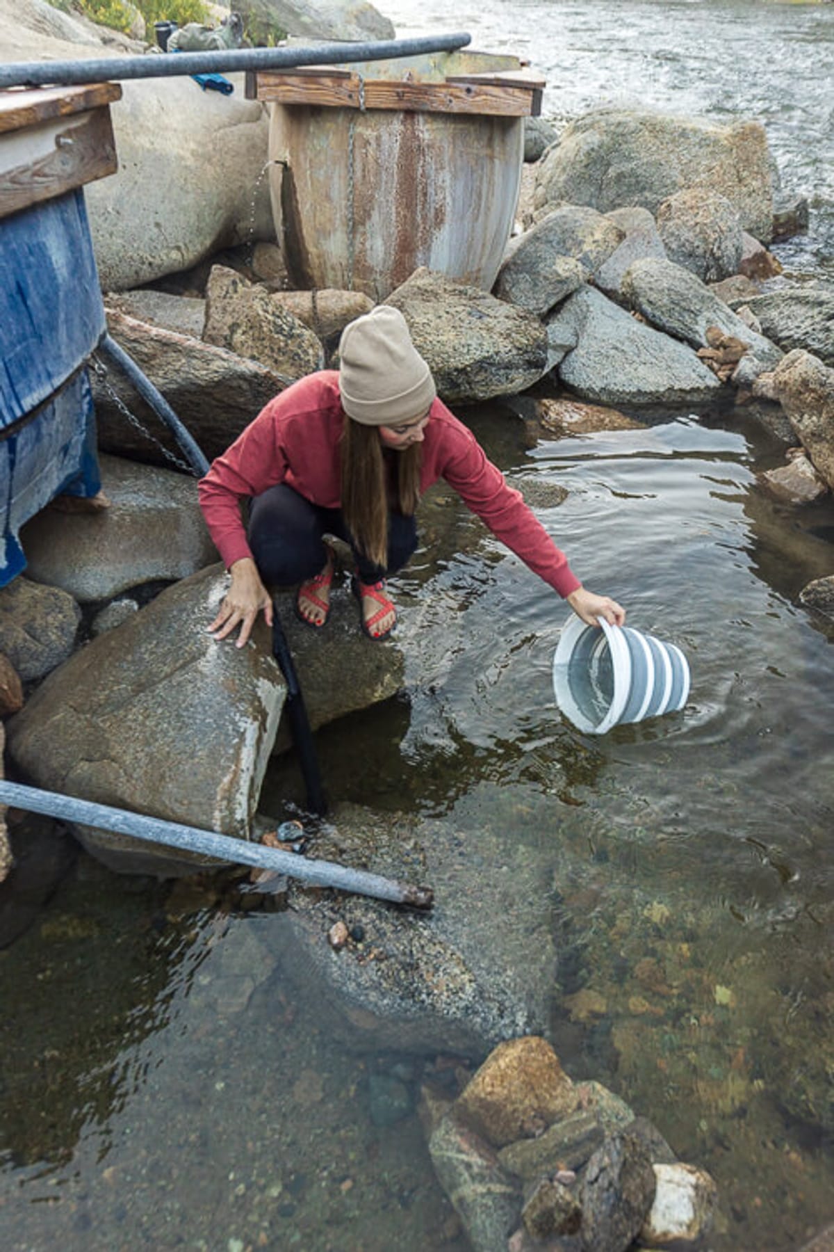 A woman bends down to fill a bucket with cold river water next to a hot spring tub.