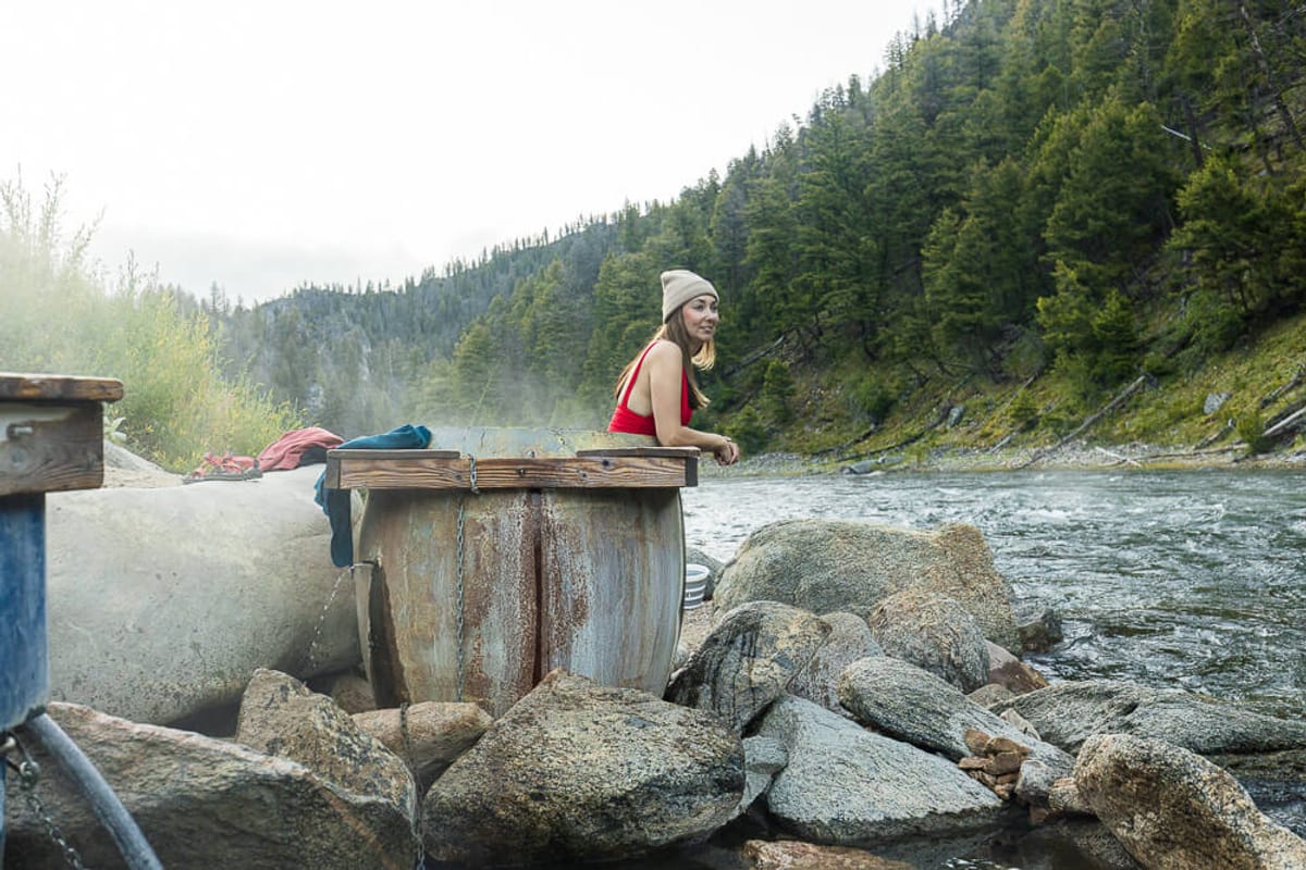 A woman in a red swimsuit and tan beanie leans over the edge of a hot spring tub and smiles looking over the adjacent river.